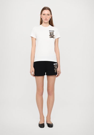 Camiseta blanca de algodón con estampado de oso y texto "MOSCHINO", combinada con pantalones cortos negros que presentan el mismo diseño de oso y logo.
