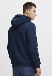 Granatowa bluza z kapturem wykonana z miękkiego materiału, o luźnym kroju, z prążkowanymi ściągaczami przy mankietach i hemie oraz z kieszenią kangurką z przodu.