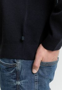 Roka v žepu modrih zbledelih hlačah, oblečen v temno teksturirano pleteno jopico z majhno črno oznako blizu roba.
