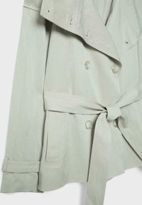 Jasnobeżowy, dwurzędowy trencz z szerokimi klapami, paskiem zawiązanym w talii, przednimi kieszeniami oraz detalem paska na mankiecie rękawa.