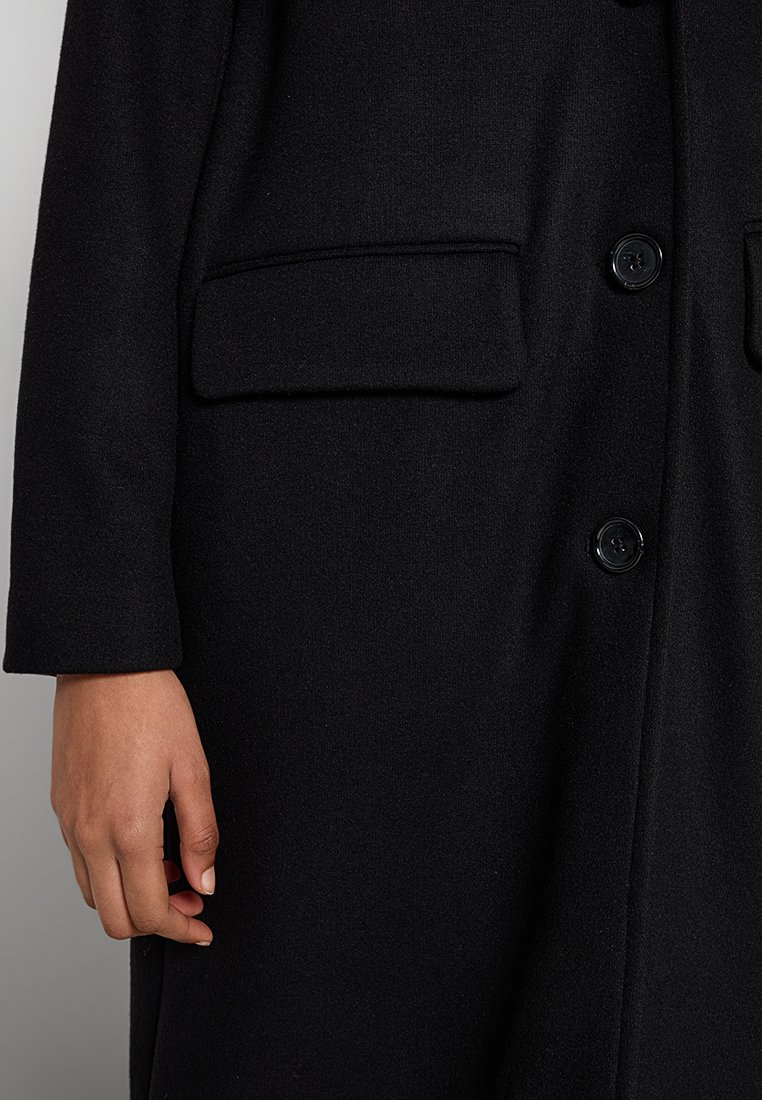 Schwarzer Mantel aus strukturiertem Stoff, mit zwei Fronttaschen und einem Knopfverschluss. Die Ärmel sind an den Handgelenken leicht tailliert.