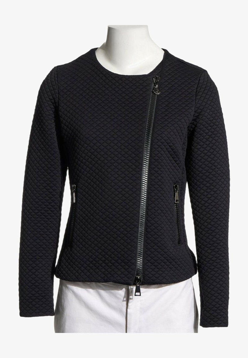Veste noire matelassée manches longues avec fermeture éclair diagonale devant et poches latérales zippées sur un buste de mannequin blanc.