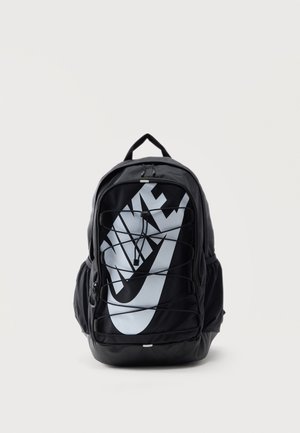 Czarny plecak Nike z dużym białym logo swoosh, elastycznymi sznurkami na froncie, bocznymi kieszeniami i górnym uchwytem na jednolitym jasnym tle.