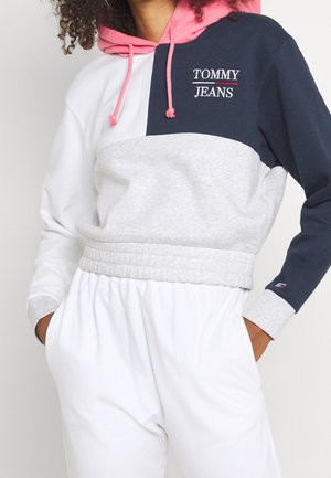 Personne portant un sweat à capuche color block avec capuche rose, manches bleu marine et blanches, milieu gris, logo "Tommy Jeans", et pantalon blanc avec les mains dans les poches.