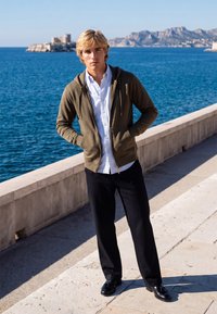 Jeune homme portant un sweat à capuche vert zippé, une chemise blanche et un pantalon noir, debout sur un chemin en pierre au bord de la mer avec un château et des montagnes en arrière-plan.