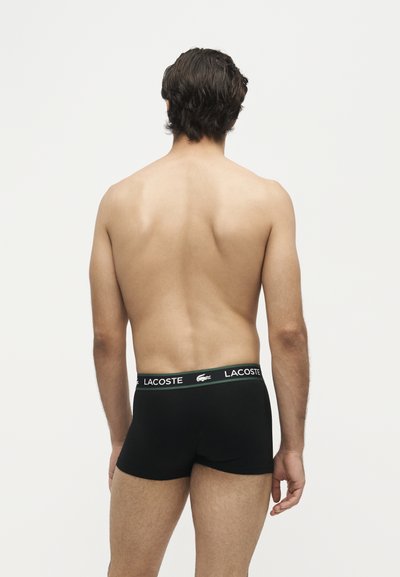 Homme aux cheveux courts et foncés, de dos, portant un boxer noir Lacoste sur un fond clair uni.