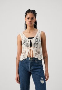 Débardeur en tricot crème avec un motif en dentelle, doté d'une fermeture devant à nouer, porté sur un haut noir, associé à un jean bleu déchiré.