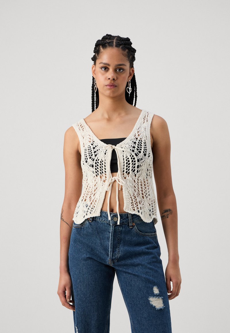 Débardeur en tricot crème avec un motif en dentelle, doté d'une fermeture devant à nouer, porté sur un haut noir, associé à un jean bleu déchiré.