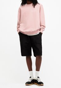 Personne portant un sweat-shirt rose AllSaints, un short noir mi-genou, des chaussettes blanches et des baskets noires, debout les mains dans les poches.