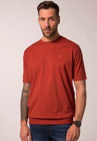 Červené bavlněné triko s kulatým výstřihem a krátkými rukávy, na hrudi s malým vyraženým logem, v kombinaci s modrými džínami a hnědými hodinkami.