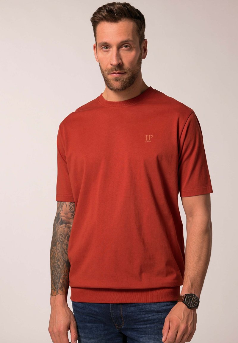 Červené bavlněné triko s kulatým výstřihem a krátkými rukávy, na hrudi s malým vyraženým logem, v kombinaci s modrými džínami a hnědými hodinkami.