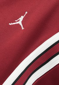 Czerwony teksturowany materiał z białym logo Jumpman oraz zakrzywionymi białymi, czarnymi i czerwonymi paskami.