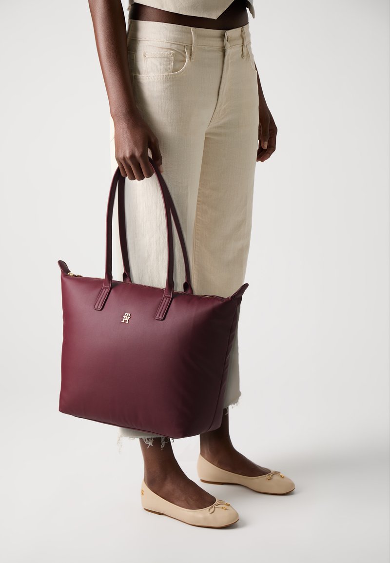 Sac fourre-tout bourgogne avec deux poignées, texture lisse ressemblant au cuir, détail logo doré, associé à un pantalon large crème et des ballerines beiges.