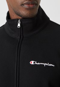 Chaqueta negra con cremallera fabricada en un tejido suave, presenta un cuello alto y el logo bordado de "Champion" en blanco y rojo. El tirador de la cremallera es de color plateado.