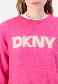 Höyhenmäinen vaaleanpunainen neulepaita, jossa on valkoinen "DKNY"-logo tekstuurilla kirjailtuna. Suunnittelu sisältää pyöreän kaula-aukon ja pitkät hihat.