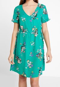 Türkisfarbend blumiges Kleid mit kurzen, gerafften Ärmeln, V-Ausschnitt, elastischem Bund und Bindedetail. Hat mehrfarbige Blumenmuster auf dem gesamten Kleid.