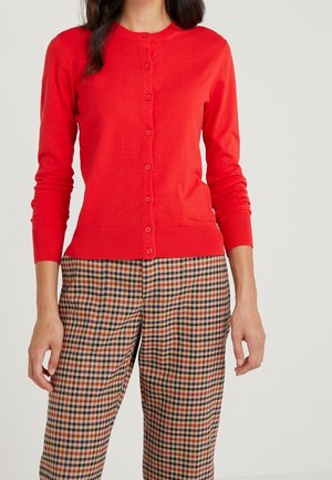 Kvinde iført en tætsiddende rød cardigan med knapper, lange ærmer og højtaljede ternede bukser i brunt, sort og rødt.