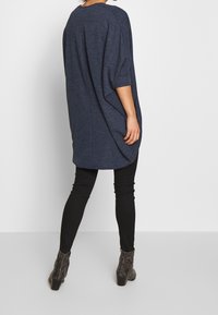 Frau trägt einen dunkelblauen übergroßen Pullover mit dreiviertel Ärmel, schwarze Leggings und graue Schlangenleder-Print-Ankle Boots und steht mit dem Rücken zur Kamera.