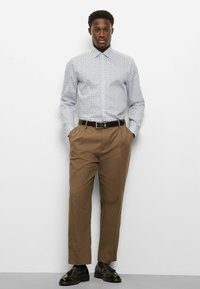 Langärmeliges, hellblau gemustertes Hemd mit spitzem Kragen, kombiniert mit braunen, plissierten Hosen und schwarzen Anzugschuhen. Brauner Ledergürtel inklusive.