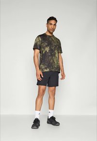 Hombre de pie, vistiendo una camiseta de hilo verde y negra, pantalones cortos negros, calcetines blancos de marca y zapatillas negras, contra un fondo liso.