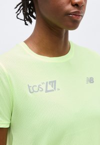 Κοντινό πλάνο ατόμου που φοράει αθλητική μπλούζα ανοιχτού πράσινου χρώματος με μικρά λογότυπα "tcs 2026" και "NB" στο στήθος.