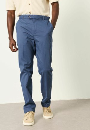 Male wearing blue tailored trousers and beige casual sneakers, standing with one hand in pocket against a plain light background.