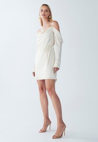 Biele off-shoulder šaty s dlhými rukávmi, stiahnutým pásom a hladkou textúrou. Nosené s nahými vysokými podpätkami.