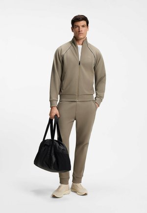 Mand iført beige tracksuit med lynlås og beige sneakers, der holder en sort træningstaske, stående foran en ensfarvet hvid baggrund.