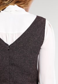 Chaleco de tweed oscuro con una espalda en V pronunciada, texturizado con manchas rosas y una cremallera oculta en la parte trasera, sobre una blusa blanca con un cuello alto.