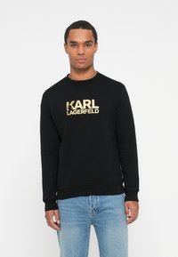 Muž v čiernej mikine s viditeľným zlatým textom "KARL LAGERFELD" a svetlomodrých džínsoch, stojaci na pozadí bielej farby.
