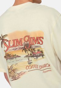 Cremefarvet bomulds t-shirt med et farverigt retro grafik med palmer, vand og en hytte-scene. Teksten lyder: "SLIM JIMS COFFEE SHACK Panama."