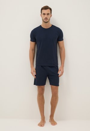 Man standing against a plain wall wearing a navy blue short-sleeve T-shirt and matching shorts, barefoot, looking forward.