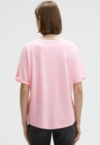 Woman with straight brown hair wears a loose light pink short-sleeve top and dark jeans, standing facing away against a plain background.