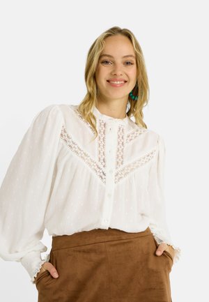 Blusa blanca de manga larga con detalles de encaje, tela texturizada, tapeta de botones y puños con volantes, combinada con una falda de ante marrón.