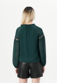 Mørkegrønn bluse med gjennomsiktige lange ermer, blonder detaljer og rynket halslinning. Kombinert med svarte faux skinnshorts. Bakside.