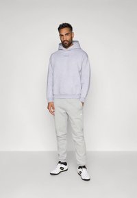 Man in een lichtgrijze hoodie, bijpassende sweatpants en witte sneakers die tegen een effen achtergrond staat.