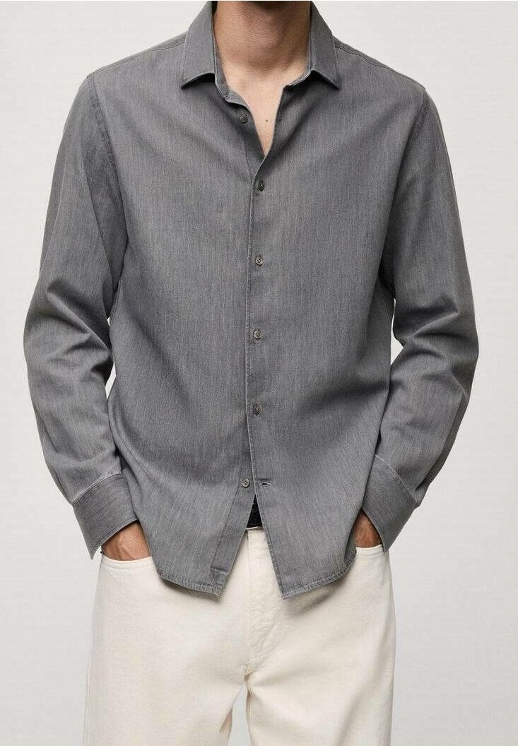 Homme portant une chemise grise à manches longues boutonnée, les mains dans les poches d'un pantalon écru, sur un fond uni.