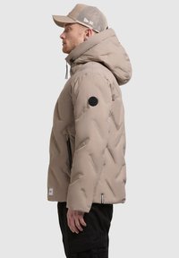 Giacca puffer beige con un motivo trapuntato strutturato, cappuccio regolabile e una toppa con logo nera. Indossata con un cappellino in rete grigio e pantaloni neri.