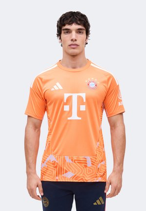 Mand står med fronten mod kameraet iført en orange FC Bayern München fodboldtrøje med hvide logoer og mønstret nederste del.