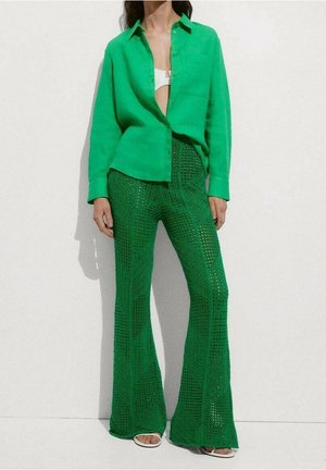 Femme portant une chemise boutonnée vert vif par-dessus un haut blanc, associée à un pantalon large texturé vert et des sandales blanches à bout ouvert, sur fond blanc.