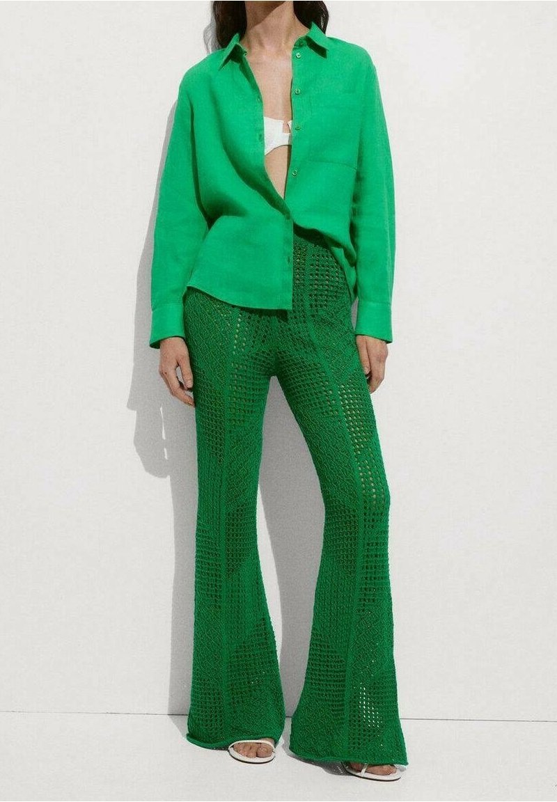 Femme portant une chemise boutonnée vert vif par-dessus un haut blanc, associée à un pantalon large texturé vert et des sandales blanches à bout ouvert, sur fond blanc.