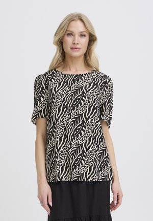 Femeie blondă purtând un top bej cu mâneci scurte, cu imprimeu abstract negru cu frunze și animale, asortat cu o fustă neagră, stând în fața unui fundal alb.