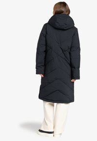 Abrigo de plumas negro con capucha, patrón de rombos, mangas largas y aberturas laterales. Lleva encima un pantalón de pierna ancha color crema.