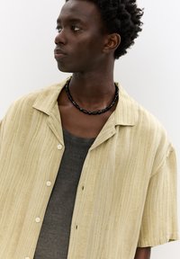 Kortærmet skjorte i lys gul med lodrette striber, lavet af blødt stof; båret over en grå tanktop og accessoriseret med en sort perlekæde.