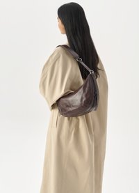 Sac à épaule en cuir marron avec une forme arrondie et une bandoulière ajustable, porté par-dessus un vêtement oversized beige clair. Texture lisse, matériel minimal.