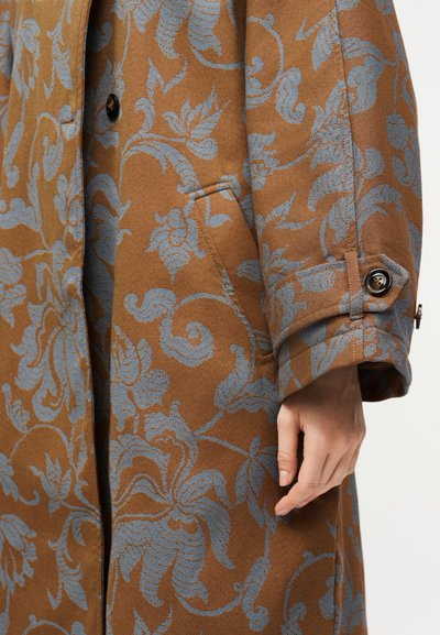 Καφέ παλτό με μπλε λουλουδάτα σχέδια, με κουμπωτό μανίκι και ένα χέρι που ακουμπά κοντά στην πλαϊνή τσέπη.