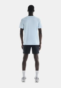 Lichtblauw sport T-shirt met korte mouwen, gecombineerd met donkerblauwe shorts. Model draagt witte sokken en hardloopschoenen, gezien vanaf de achterkant.