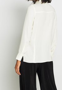 Blouse à manches longues couleur crème avec poignets boutonnés, texture lisse, détail à l'empiècement dos, associée à un pantalon large noir texturé.