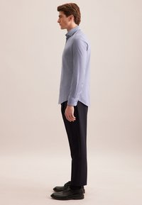 Jonge man die in profiel staat, gekleed in een lichtblauw overhemd, zwarte broek en zwarte nette schoenen tegen een beige achtergrond.