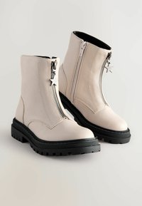 Bottes à chevilles crème avec semelles noires épaisses, zips avant avec tirettes en forme d'étoile et détails de zip sur les côtés.